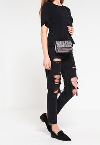 Nainen, joka on pukenut ylleen mustan kuvioidun lyhythihaisen paidan, voimakkaasti revityt mustat farkut, mustat liukuva kengät sekä pienen metallisen ristiin kantettavan laukun koristeilla.