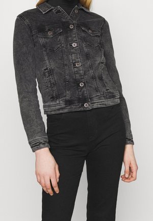 Person iført en tætsiddende sort acid-wash denimjakke med knaplukninger og sorte højhælede bukser mod en ensfarvet baggrund.