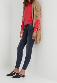 Cárdigan beige de punto superpuesto a un suéter rojo, combinado con jeans ajustados oscuros y mule negros con detalles de tachuelas blancas.