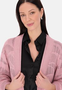 Cardigan a maglia rosa chiaro con apertura frontale, caratterizzato da motivi testurizzati e bordi a coste, indossato sopra una camicia nera con bottoni.