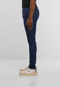 Vaqueros de mezclilla en azul oscuro con corte ajustado, que cuentan con cinco bolsillos y costuras sutiles. Combinados con zapatillas crema y beige.