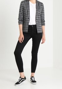 Černo-bílý kostkovaný blazer přes bílé tričko, spárovaný s černými slim džínami a černými teniskami s bílou podrážkou.