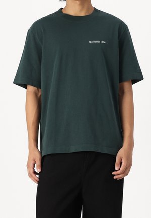 Personne portant un t-shirt à manches courtes vert foncé avec le texte "Abercrombie | NYC" sur la poitrine gauche, associé à un pantalon noir.