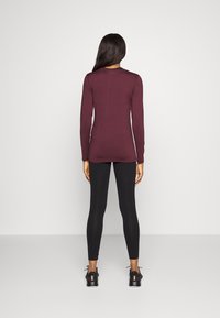 Bordeaux langermet topp med en tettsittende design, kombinert med svarte leggings. Modellen har på seg svarte joggesko med rosa detaljer.