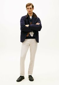 Marineblauwe jas met bruine kraag, witte blouse, gemusterde trui in bruin, wit en marineblauw, lichtbeige broek en zwarte schoenen.
