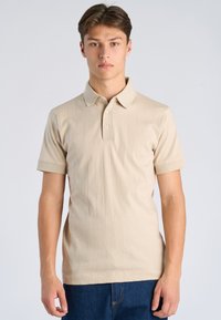 Junger Mann steht, trägt ein beiges Polohemd mit kurzen Ärmeln und dezenten vertikalen Streifen sowie blaue Jeans, vor einem schlichten, hellen Hintergrund.