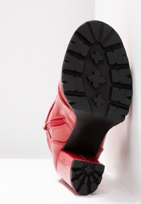 Bottine rouge à talon carré, semelle texturée avec motif de crampons en forme de croix, et fermeture éclair latérale, positionnée en angle sur un fond blanc.