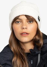 Witte fleece beanie met een opgerolde rand en een zwart logo-label. Het model draagt een donkere jas eronder, wat een gladde textuur laat zien.