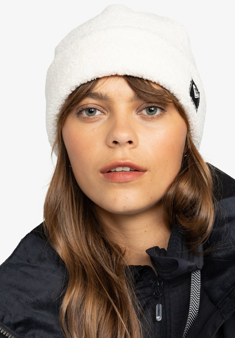 Witte fleece beanie met een opgerolde rand en een zwart logo-label. Het model draagt een donkere jas eronder, wat een gladde textuur laat zien.