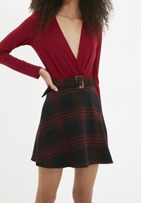 Body rouge à manches longues avec un décolleté en V profond, associé à une jupe à carreaux noir et rouge avec une large ceinture à boucle rectangulaire.