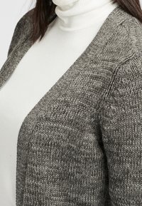 Grauer gestrickter Cardigan mit offenem Front und strukturiertem Stoff. Über einem weißen Rollkragenpullover getragen, hebt er sein gemütliches Design und die neutrale Farbe hervor.