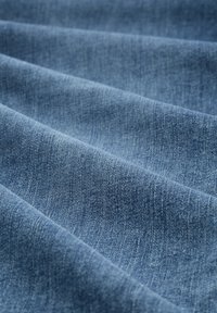 Denim tkanina svijetloplave boje s glatkom teksturom, koja sadrži suptilno izblijeđivanje i klasični tkanina uzorak. Obješena u nježnim valovima.