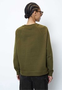 Persona con cabello trenzado y gafas, vistiendo un sudadera verde oliva y pantalones negros, de espaldas contra un fondo blanco liso.