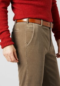 Corduroy broek in een gedempte bruine kleur met verticale strepen, voorzien van zijzakken en een bruine leren riem, gedragen met een rode gebreide trui.