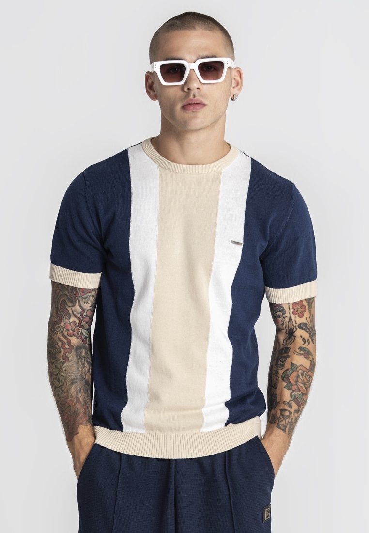 Hombre con cabello corto y brazos tatuados que lleva gafas de sol blancas cuadradas y una camisa de manga corta a rayas en azul marino, beige y blanco, con las manos en los bolsillos.