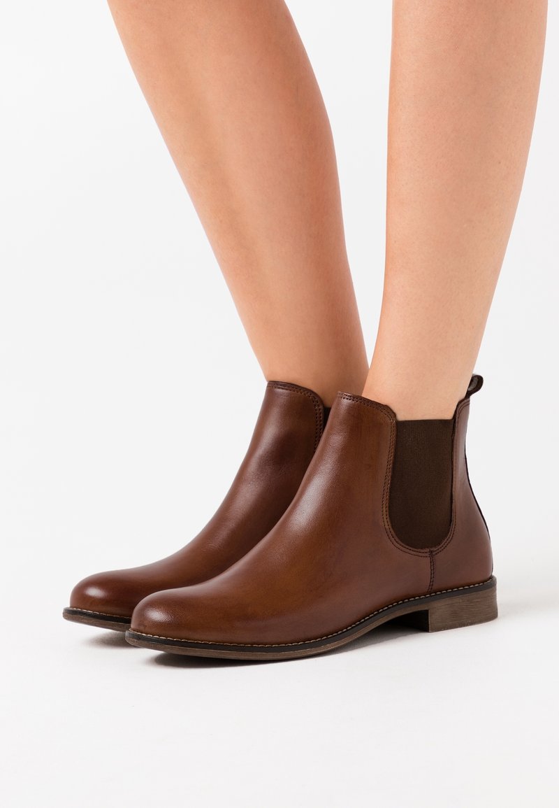 Bottines Chelsea en cuir marron avec des panneaux élastiques sur les côtés, portées sur des jambes nues, présentées sur un fond blanc.