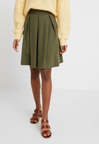 Olivgrön plisserad kjol i mjukt tyg, med utsvängd nederdel och elastisk midja, kombinerad med bruna sandaler med remmar.