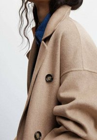 Abrigo de mezcla de lana beige con cuello de muesca, cuatro grandes botones y una silueta suavemente drapeada, llevado sobre una camisa de mezclilla azul.