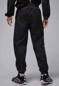 Black track pants made of lightweight material, featuring elastic cuffs and side stripes with a dotted pattern. Worn with black athletic shoes.