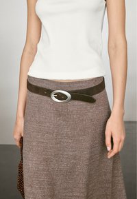 Bruine leren riem met een ronde zilveren gesp, gedragen over een gebreide bruine rok en een witte aansluitende top. Soepele textuur en strak ontwerp.