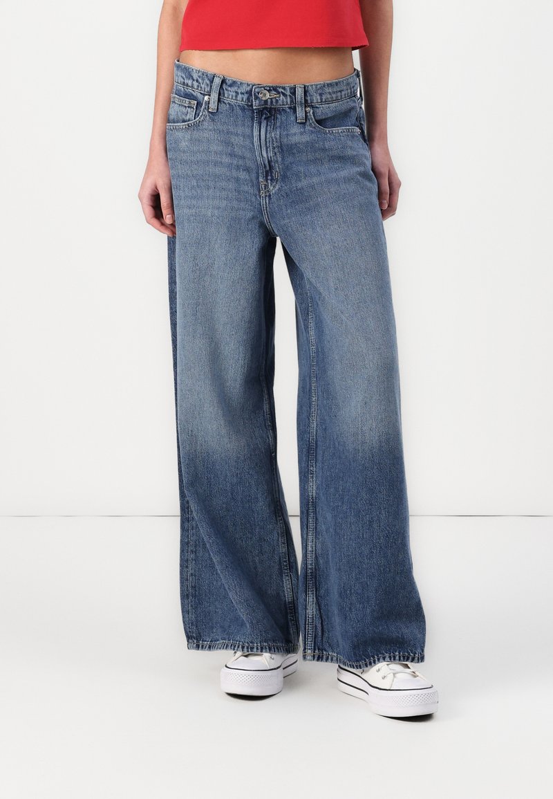 Wide-leg blue denim jeans with a faded wash, featuring a button and zip fly, five pockets, and frayed hem details.