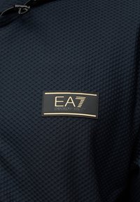 Υφασμα σε ναυτικό μπλε με υφή και ανάγλυφο σχέδιο, με ένθετη ετικέτα λογότυπου EA7 Emporio Armani σε μαύρο και χρυσό να ξεχωρίζει εμφανώς.