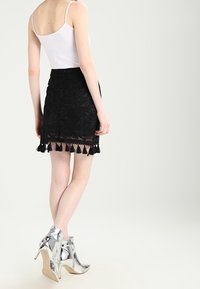 Černá krajková mini sukně s vlnkovaným lemem a třásněmi, kombinována s bílým topem a stříbrnými kotníkovými botami. Hladká textura a přiléhavý střih.