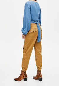 Blouse bleu clair nouée dans le dos, pantalon cargo beige avec poches latérales, et bottines marron à motif serpent avec talon bloc.