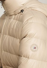 Beżowa kurtka puchowa z pikowanym wzorem, wyposażona w wysoki kołnierz, zamek błyskawiczny i naszywkę z logo na lewym ramieniu. Gładka tekstura.
