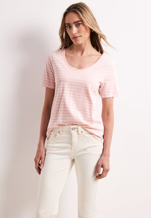 Frau trägt ein hellrosa gestreiftes Kurzarmhemd, das in hochgeschnittene, off-white Jeans gesteckt ist, und steht vor einem einfarbigen Hintergrund.