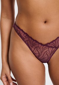 String violet en dentelle transparente avec bords festonnés et détails à motif floral, conçu pour une coupe près du corps. Texture douce et délicate.