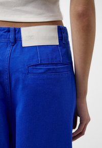 Blå denimbyxor med en bakficka och en vit läderlogg vid midjan. Texturen är slät med ett synligt vävt mönster.