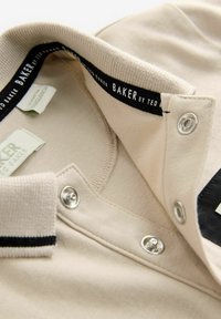 Bež polo majica s crnom obrubom na ovratniku, metalnim kopčama na dugmad i oznakom "BAKER by Ted Baker" unutar ovratnika. Mekani pamuk.