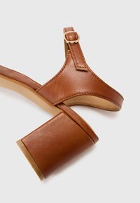 Brune lærreim sandal med hæl som har en firkantet blokkhels og en gullfarget spenne på reimen. Glatt tekstur med sømdetaljer.