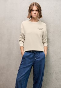 Felpa beige con il testo "CITY CLUB" sul petto, abbinata a pantaloni in denim blu scuro larghi con strisce laterali e vita con cordino regolabile.