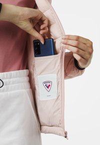 Ljust rosa isolerad jacka med en ficka för telefon. Har en vit etikett med röd och blå logotyp samt en dragkedja. Hand som sätter in en telefon i fickan.