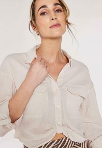 Helles beige Button-Up-Hemd mit zwei Taschen und einem geknoteten Frontdetail, aus strukturierter Stoff gefertigt. Weiches, lässiges Design mit hochgerollten Ärmeln.