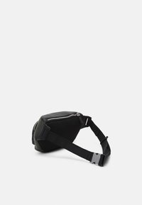 Musta vyötärölaukku, jossa on vetoketjullisia lokeroita ja säädettävä hihna, jossa on metallinen solki, asetettuna valkoiselle pinnalle.