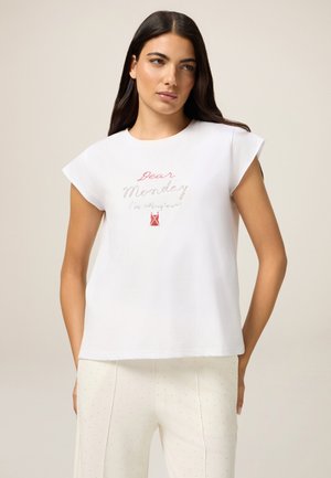 T-shirt bianco in cotone con maniche corte a sbuffo, decorato con un testo ricamato colorato "Dear Monday" e una piccola grafica rossa. Indossato con pantaloni chiari.