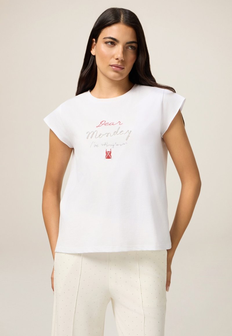 Camiseta de algodón blanca con mangas cortas tipo cap, que presenta un colorido texto bordado "Querido Lunes" y un pequeño gráfico rojo. Combinada con pantalones claros.