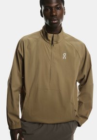 Braune leichte Jacke mit hohem Kragen, halbseitigem Reißverschluss und Fronttasche. Mit Logo auf der Brust und elastischen Bündchen.