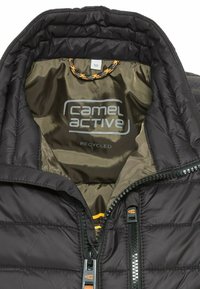 Chaqueta negra acolchada con cremallera abierta que muestra un forro de satén verde, de la marca "camel active" y etiqueta de tamaño "50".