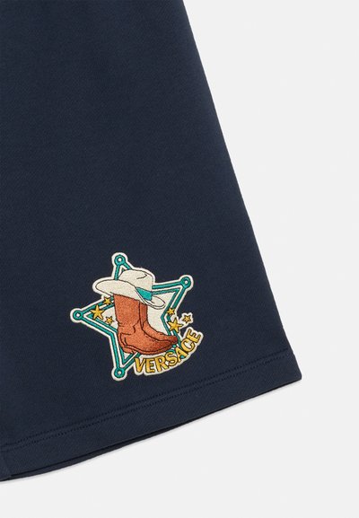 Υφασμα σε ναυτικό μπλε με κεντημένη μπαλώματα που δείχνει ένα καφέ κάουμποϋ μπότες, λευκό καπέλο, μπλε αστέρι, κίτρινα αστέρια και το κείμενο "Versace".