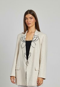 Donna con capelli castani lisci che indossa un blazer beige con ricami floreali neri sopra una maglietta nera, contro uno sfondo grigio semplice.