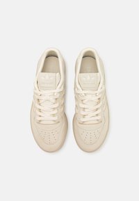 Beige Adidas sneakers with a smooth leather upper, round toe, perforated detailing, flat laces, and three signature stripes on the sides.