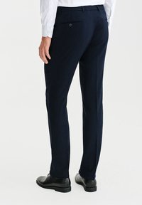 Granatowe spodnie garniturowe z gładkiego materiału, z prostą nogawką, tylną kieszenią z guzikiem, noszone z czarnymi butami.