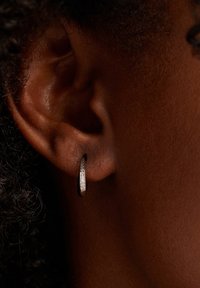 Boucle d'oreille en argent avec une surface texturée, dotée d'une rangée de petites pierres claires d'un côté, mise en valeur sur une peau foncée et des cheveux bouclés.