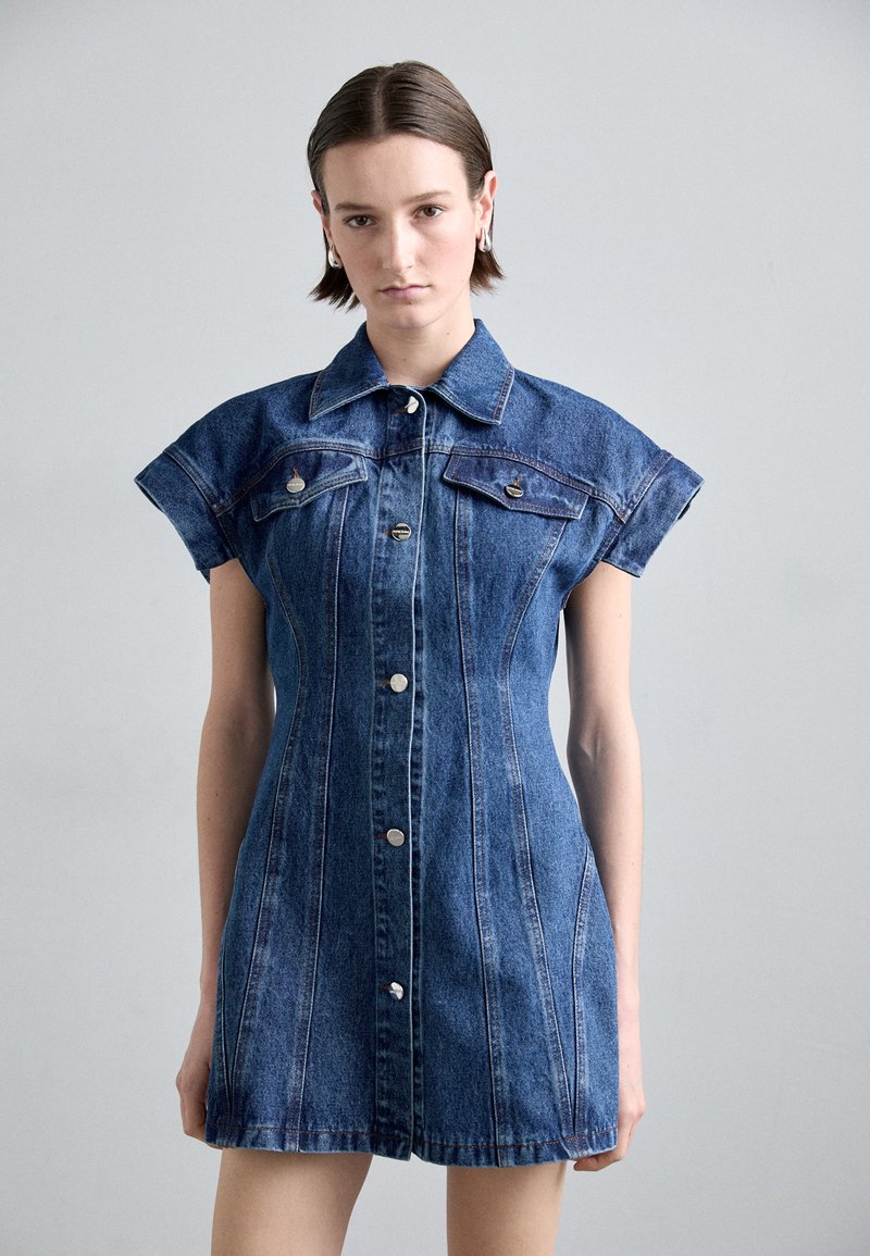 Rochie din denim cu o siluetă ajustată, maneci scurte, buzunare frontale și detalii cu nasturi argintii. Culoare albastră medie cu linii de cusătură vizibile.
