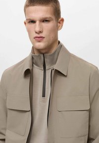 Beige jas met een hoge kraag, twee voorzakken en een contrasterende rits. Draagt bovenop een lichtbeige pullover met een kwart rits.