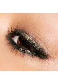 Gros plan sur un œil avec un iris marron foncé, souligné par un fard à paupières vert profond scintillant et de longs cils noirs recourbés. La peau est lisse.